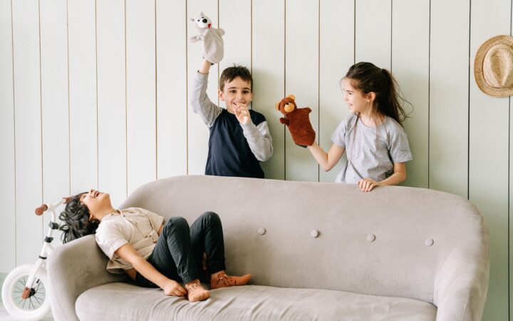 Trois enfants jouent avec des marionnettes derrière un canapé dans un intérieur lumineux