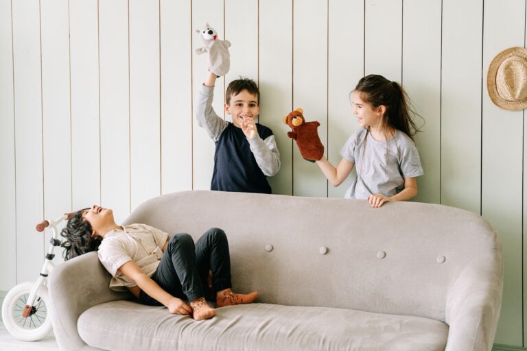 Trois enfants jouent avec des marionnettes derrière un canapé dans un intérieur lumineux