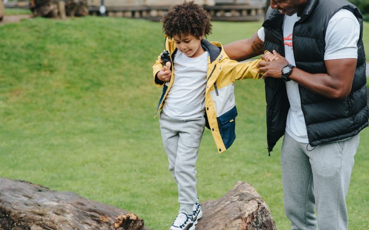 Un parent aide son enfant à garder l’équilibre en marchant sur un tronc d’arbre dans un parc.