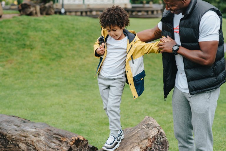 Un parent aide son enfant à garder l’équilibre en marchant sur un tronc d’arbre dans un parc.